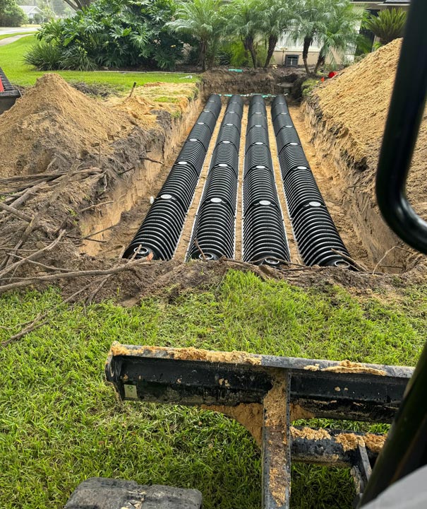 New drain field with four rows of black pipe in an open trench, seen from the excavator